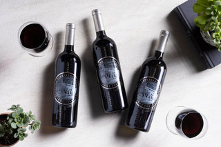 Three wine bottles with labels on a light surface, accompanied by two glasses of red wine.